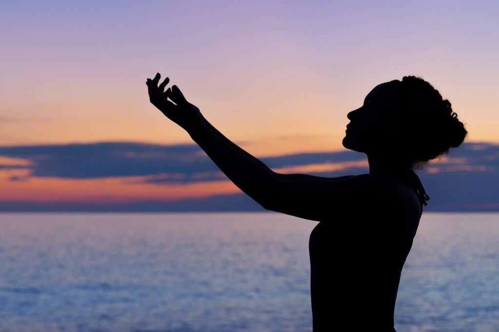 Young woman raising hands to the sky at sunrise, investing in her body, mind, and spirit bank accounts.